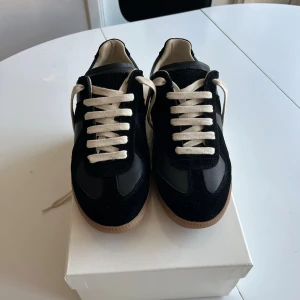Maison Margiela Gats Black Gum - Stilrena svarta gats med beige gummisula och vita snören. Ovandelen är i en mix av mocka och slätt läder, vilket ger en snygg kontrast. Allmänt sköna skor som passar till allt. Perfekta för dig som gillar minimalistisk och tidlös stil. Säljer eftersom de var för små på mig. Oanvända. Dustbag + box medföljer. Hör av dig vid minsta fundering!