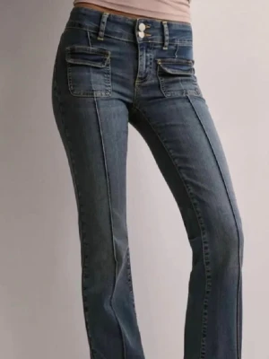 Blå jeans med fickdetaljer från Nelly - Säljer ett par blå bootcut jeans från Nelly med dubbla knappar i midjan och  fickor   framtill. Inte särskilt använda, men har en liten defekt vid ett av ”bält hållarna” se bild 7 