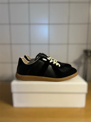 Svarta Maison Margiela Replica sneakers - Svarta Maison Margiela Replica sneakers med ovandel i mocka och läder, vita snören och beige innersula. Passar mellan 40-42