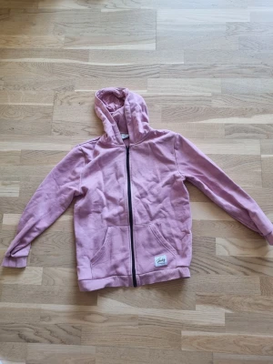 Rosa hoodie med dragkedja från Tandy - Säljer en rosa hoodie med dragkedja framtill och två stora fickor. Hoodien har en klassisk huva och är gjord i mjukt bomullsmaterial. Perfekt för chill dagar eller när du vill ha en avslappnad stil. Från stadium outlet!