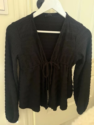 Svart ribbad kofta med knytband - Trendig svart ribbad kofta med långa ärmar och knytband framtill. Koftan har en djup V-ringning och är tillverkad i ett stretchigt, våfflat material som ger en snygg struktur. 