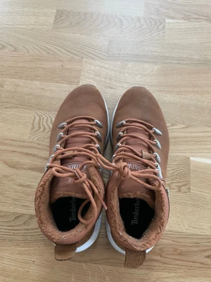 Bruna Timberland kängor i mocka - Snygga bruna kängor från Timberland i mocka med vita och svarta sulor. Kängorna har metallhål för snörning, vadderad krage och Timberland-logga på sidan. Perfekta för höst och vinter med sin robusta design och sköna innersula.