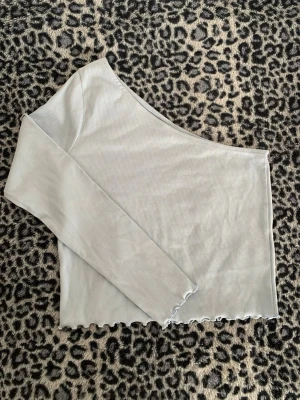 Ljusblå oneshoulder topp Gina Tricot - Trendig ljusblå oneshoulder topp från Gina Tricot i ribbad bomullsmix. Toppen har lång ärm på ena sidan och vågig kant nedtill för en cool detalj. Perfekt för dig som vill sticka ut lite extra. I storlek XL men passar även S-M