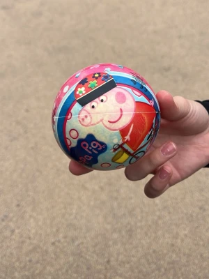 Peppa Pig boll med färgglatt motiv - Säljer en rund boll med Peppa Pig-motiv i rosa, blått och rött. Bollen har tecknade blommor och Peppa Pig i röd klänning på framsidan. Perfekt för lek utomhus eller inomhus. Tillverkad i plast.