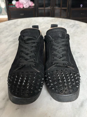 Svarta sneakers med nitar - Svarta sneakers från Christian Louboutin i mocka med svarta nitar över tån och svarta skosnören. Låg modell med rund tå och diskreta detaljer på sidorna. Insidan är beige och sulan är svart. Perfekta för dig som vill sticka ut med en edgy stil. Men de är lite slitna i insidan samt så saknas det 2 spikar, inget som märks. Då du har på dig skon och inte visar hur de ser ut inuti för allmänheten😅Spikarna, en är av längst ner på sidan utav skon och andra är längst fram. 