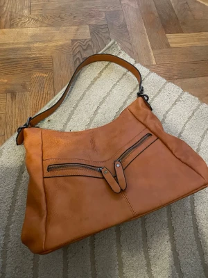 Brun axelväska med dubbla dragkedjor - Snygg axelväska i brunt läder med två diagonala dragkedjor framtill och bred axelrem. Väskan har en mjuk, lite slouchy form och detaljer i svart metall. Perfekt för dig som vill ha en stilren men ändå cool väska till vardagslooken. Superfin och inga defekter alls❤️ Använd gärna köp nu ❤️