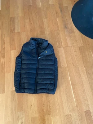 Lyle & scott väst - Snygg svart dunväst från Lyle & Scott med gul logga på bröstet. Västen har quiltad design, dragkedja framtill och står upp lite i kragen. Perfekt att slänga över en hoodie eller tröja för extra värme. Materialet är lätt och smidigt.