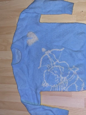 Blå stickad tröja med amor-motiv - Ljusblå stickad tröja med vitt motiv av en amor med pilbåge och ett hjärta på framsidan. Tröjan har rund halsringning och långa ärmar. Perfekt för dig som gillar gulliga och unika plagg med lite retrovibe.
