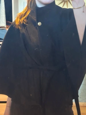 Svart cape-kappa med bälte - Stilren svart cape-kappa med vid ärm och hög krage. Kappan har ett dekorativt guldknapp vid halsen och knyts i midjan med ett matchande bälte. Perfekt för dig som vill ha en elegant och trendig look. Materialet ser ut att vara ullblandning.😍🩷 
