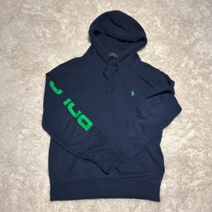 Marinblå hoodie Polo Ralph Lauren - Snygg marinblå hoodie från Polo Ralph Lauren med grön logga på bröstet och stort grönt tryck med texten POLO RALPH LAUREN längs ena ärmen. Klassisk huva med snörning och känguruficka framtill. Perfekt för en chill och sportig look.Skicket är bra och säljs pga för liten