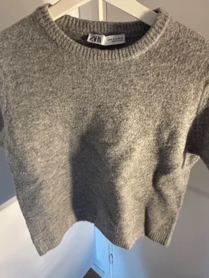 Grå stickad tröja från Zara - Mysig grå stickad tröja från Zara med rund halsringning och ribbade muddar. Tröjan har en enkel, klassisk design och är tillverkad i ett mjukt material som passar perfekt för kyligare dagar.