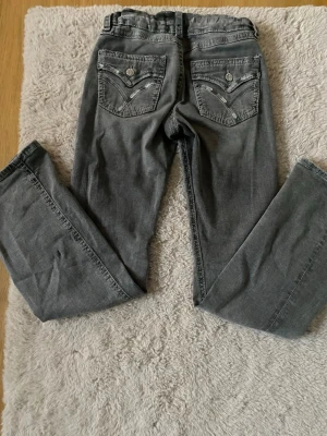 Grå jeans med detaljerade bakfickor - Säljer ett par grå jeans med snygga detaljer på bakfickorna, inklusive knappar och dekorativa sömmar. Jeansen har en rak passform och klassisk femficksdesign. Perfekta för dig som vill ha en avslappnad men ändå stilren look.