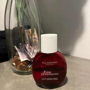 Clarins Eau Dynamisante parfym - En rund parfymflaska från Clarins Aroma-serie i rödtonat glas med vit kork. Flaskan har elegant design och texten 'Eau Dynamisante' på framsidan. Perfekt för dig som gillar fräscha och energigivande dofter. Hälften kvar
