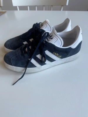 Adidas Gazelle Mörkblå - Mörkblå adidas gazelle skor med vita detaljer. Storlek 39 1/3, använda ett fåtal gånger men syns fof på det vita att amn gått med dom utomhus men det går att städa rent om man vill få dom helt vita (se bild)💞