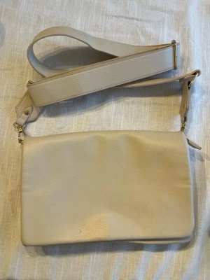 Beige axelväska med bred rem - Stilren beige axelväska i mjukt konstläder med bred justerbar axelrem och guldfärgade metalldetaljer. Väskan har enkel, rektangulär form och en diskret ficka på baksidan. Perfekt för dig som gillar minimalistisk och clean stil.