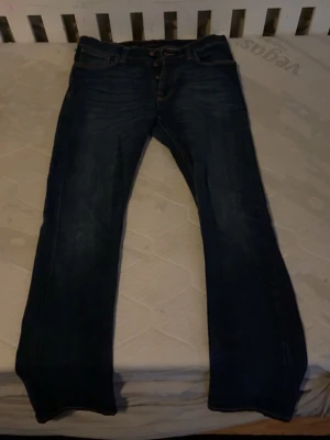 Mörkblå jeans från Nudie Jeans - Säljer ett par mörkblå jeans från Nudie Jeans med klassisk femficksmodell och raka ben. Jeansen har orangea sömmar och knäppning fram. Perfekta för dig som gillar en tidlös och clean look.