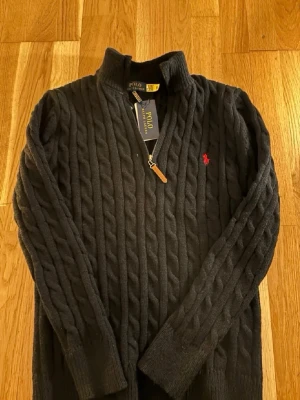 Svart kabelstickad tröja Polo Ralph Lauren - Kabelstickad svart tröja från Polo Ralph Lauren med halv dragkedja och röd broderad logga på bröstet. Tröjan har hög krage och ribbade muddar. Perfekt för dig som gillar klassisk och stilren design.