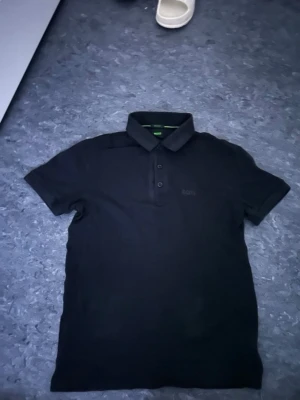 Svart polotopp från BOSS - Säljer en svart polotopp från BOSS med klassisk krage och knappar framtill. T-shirten har korta ärmar och är tillverkad i bomull, vilket gör den både bekväm och stilren. Perfekt för dig som gillar en enkel och clean look.