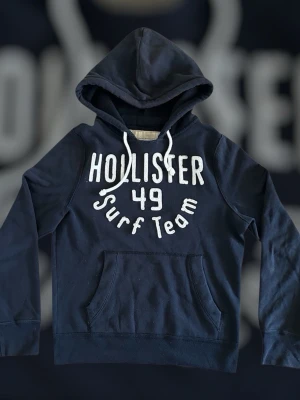 Mörkblå hoodie från Hollister - Mörkblå hoodie från Hollister med vit text 'Hollister 49 Surf Team' på bröstet. Klassisk känguruficka framtill och justerbar huva med dragsko. Perfekt för en avslappnad och sportig stil.