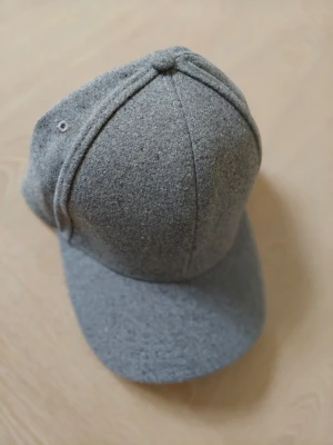 Varm grå keps i ullmix Polyester  - Snygg grå keps i mjuk ullmix med klassisk böjd skärm och sexpanelers design. Kepsen har minimalistisk look utan synligt märke och passar perfekt till en clean och stilren outfit. Materialet ger en lite lyxigare känsla och funkar året runt.