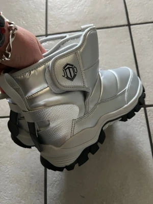 Silvriga moon boots från JTO storlek 38 - Säljer ett par silvriga moon boots från JTO med chunky svart sula och vadderad ovandel. Bootsens material är glansigt och quiltat, med en cool logga på sidan. Perfekta för vinterns kalla dagar och snöiga äventyr.