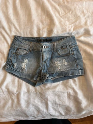 Blå denimshorts från ONLY - Snygga blå denimshorts från ONLY med slitna detaljer framtill och uppvikta benslut. Klassisk femficksmodell med knapp och dragkedja. Perfekta för varma dagar och enkel att matcha med olika toppar.