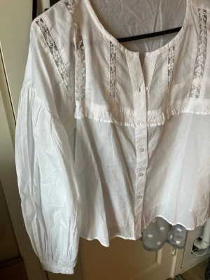 Vit blus med spetsdetaljer - Säljer en vit blus med knappar framtill och dekorativa spetsdetaljer på bröstet. Blusen har en rund halsringning och långa ärmar med lätt puff. Materialet är tunt och luftigt, perfekt för en stilren look. Säljer pga köpte fel storlek så den är aldrig använd🥰
