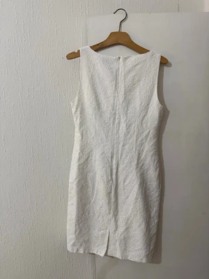 Vit kort klänning från KappAhl - Elegant vit kort klänning från KappAhl med vackert strukturerat mönster i tyget. Klänningen är ärmlös och har en rak, figurnära passform samt dragkedja i ryggen. Perfekt för dig som gillar stilrena och klassiska plagg.