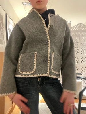 Grå cardiganjacka från ryvls  - PRIS KAN DISKUTERAS. Super fin grå cardiganjacka med dekorativa vita kanter och stora knappar framtill. Jackan har två framfickor och är tillverkad i ett tjockare ull-liknande material. Rak passform och långärmad, perfekt för våren. Originalpriset var 600kr jag är 162 cm och den passar 