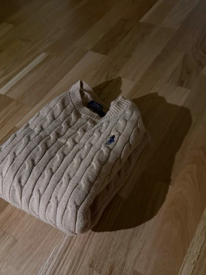 Beige kabelstickad tröja Polo Ralph Lauren - Kabelstickad tröja i beige från Polo Ralph Lauren med klassisk blå broderad logga på bröstet. Tröjan har rund halsringning och ribbade muddar. Perfekt för dig som gillar tidlös och clean stil. Materialet känns mjukt och håller värmen.