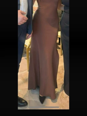 Brun långklänning från Mango - Säljer en elegant brun långklänning från Mango i modellen LAZARO. Klänningen har en figurnära passform och är ärmlös med en stilren, minimalistisk design. Perfekt för dig som gillar en klassisk och tidlös look.