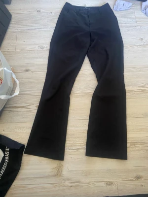 Svarta kostymbyxor med utsvängda ben - Säljer ett par svarta kostymbyxor med utsvängda ben och klassisk passform. Byxorna har hällor för skärp, diskreta bakfickor och stängs med knapp och dragkedja. Perfekta för en stilren look till vardags eller mer uppklätt. Materialet känns mjukt och följsamt.