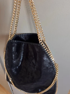 Svart handväska med guldkedjor - Snygg svart handväska i glansigt material med dekorativa guldfärgade kedjor som axelband och runt kanterna. 
