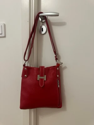 Röd axelväska med silverdetaljer i äkta läder - Snygg röd axelväska i äkta läder med justerbar axelrem och silverfärgade metalldetaljer. Väskan har en dekorativ spänndetalj framtill och en dragkedjeficka på framsidan. Perfekt för dig som vill ha en färgklick till din outfit.