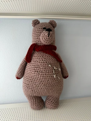 Handgjord virkad nallebjörn med halsduk och berlocker - Denna handgjorda virkade nallebjörn är en mjuk leksak. Nallebjörnen har en röd halsduk och är dekorerad med små berlocker.