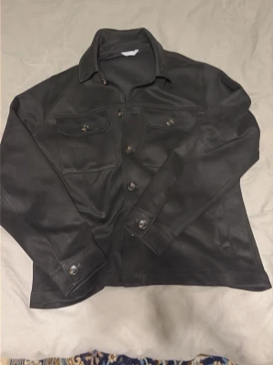 Svart cardiganjacka i skinnimitation - Snygg svart cardiganjacka i skinnimitation med klassisk krage och knappar framtill. Två bröstfickor med lock och knapp samt knappar vid ärmsluten. Jackan har en rak passform och ger en cool, stilren vibe. Väldigt bra skick och skön material. Priset kan diskuteras!!!!!