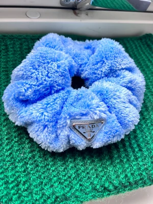 Ljusblå fluffig scrunchie från Prada - Supersöt och fluffig scrunchie i ljusblå plysch från Prada. Den har en ikonisk metallbricka med logotypen framtill och är perfekt för att ge din frisyr en lyxig touch. Materialet är mjukt och ger en trendig look. Har själv aldrig använt den, och säljer vidare den.