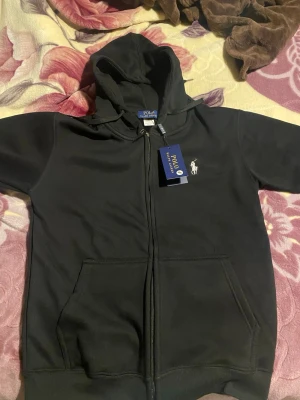  Polo Ralph Lauren - Svart hoodie från Polo Ralph Lauren med dragkedja framtill och klassisk logga broderad på bröstet. Hoodien har kängurufickor och ribbade muddar. Tillverkad i mjukt bomullsmaterial och har en skön huva. Perfekt för en avslappnad och stilren look.