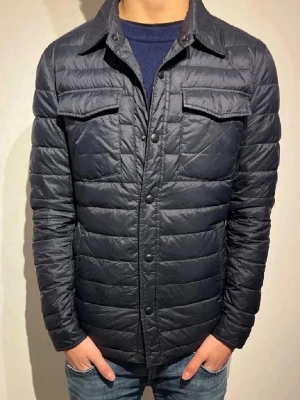 Marinblå pufferjacka med fickor - Stilren blå pufferjacka med krage och tryckknappar framtill. Två bröstfickor med lock och två sidofickor. Quiltad design som ger en modern look. 