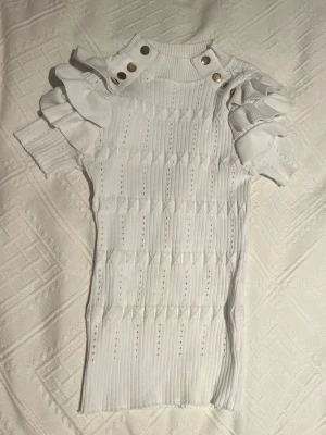 Vit ribbad topp med volangärmar - Supersnygg vit topp med ribbad struktur och dekorativa guldknappar på axlarna. Puffiga volangärmar ger en trendig look och toppen har en hög hals.