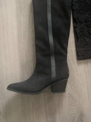 Svarta knähöga stövlar med klack - Snygga svarta knähöga stövlar i mockaimitation med bred klack och spetsig tå. Dekorativ rem i skinnimitation längs skaftet och dragkedja på insidan för enkel på- och avtagning. Perfekta för att lyfta vilken outfit som helst. Märket är Taimo, använda ett fåtal gånger då de är lite små 