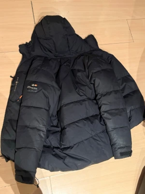 Svart dunjacka Sail Racing - Svart dunjacka från Sail Racing med Sail Antarctica Expedition-märke på bröstet. Jackan har en stor huva, kraftiga paneler och dragkedja med orange detaljer. Perfekt för kalla vinterdagar och riktigt varm.