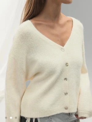 Vit kofta - Mysig krämvit kofta med v-ringning och pärlemorknappar. Koftan har en lös passform, långa ärmar och är stickad i ett fluffigt material som känns lyxigt och mjukt. Perfekt att slänga över en topp för en chill look. Storlek L men sitter som S