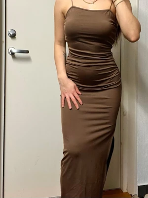 Brun figurnära maxiklänning - Säljer en brun, figurnära maxiklänning med tunna axelband och öppen rygg. Klänningen har en slits på sidan och är gjord i ett stretchigt, glansigt material som sitter snyggt mot kroppen. 