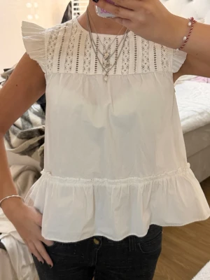 Vit ärmlös topp med spetsdetaljer - Söt vit topp med volangkant nedtill och spetsdetaljer upptill. Toppen är ärmlös med små volangärmar och har en luftig passform. Perfekt till sommaren och enkel att styla med smycken eller jeans.