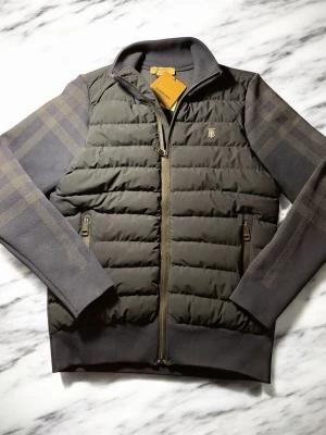 Svart pufferjacka från Burberry - Snygg svart pufferjacka från Burberry med quiltad front, ribbade muddar och dragkedja. Ärmarna har diskret rutmönster och jackan har två fickor med dragkedja. Ikonisk Burberry-logga på bröstet och hög krage för extra stil.