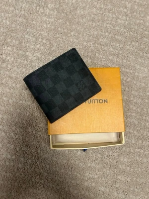 Louis Vuitton svart rutmönstrad plånbok - Snygg svart plånbok från Louis Vuitton med klassiskt rutmönster i canvas. Plånboken har en stilren och kompakt design med plats för kort och sedlar. Perfekt accessoar för dig som gillar exklusiva detaljer och tidlös stil.