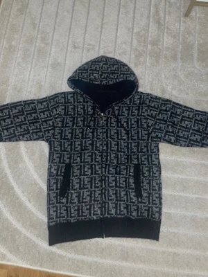 Svartgrå hoodie med mönster - Säljer en svartgrå hoodie med dragkedja och huva. Hoodien har ett fett grafiskt mönster över hela plagget och svarta muddar vid ärmar och nederkant. Två fickor framtill och mjukt material som känns skönt mot huden. Perfekt för dig som gillar streetwear vibes.