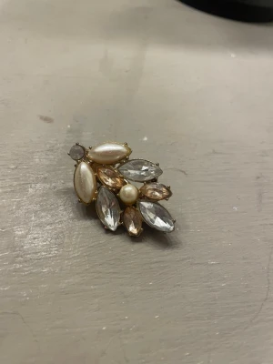 Guldig hårklämma med pärlor och stenar - Snygg hårklämma i guldmetall dekorerad med vita pärlor och glittrande stenar i olika former och storlekar. Perfekt accessoar för att ge frisyren extra bling och en elegant touch. Klämman har ett stabilt metallfäste på baksidan.