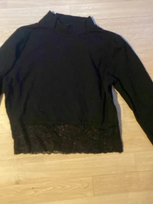 Svart croppad långärmad tröja med spets - Svart långärmad tröja med hög krage och snygg spetsdetalj längst ner. Perfekt för dig som gillar en enkel men ändå trendig look. Tröjan har en rak passform och är tillverkad i mjukt material.